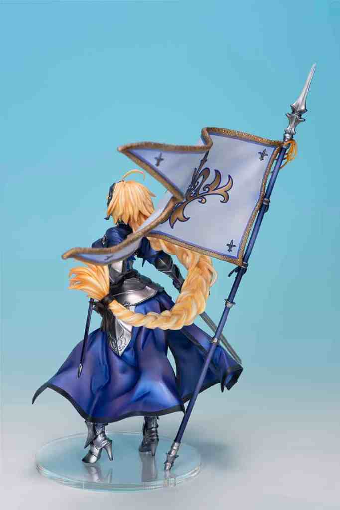 A detailed figure of Jeanne d'Arc from Fate/Grand Order, showcasing her in a blue outfit holding a flag, with an intricate hairstyle and a dynamic pose against a light blue background.