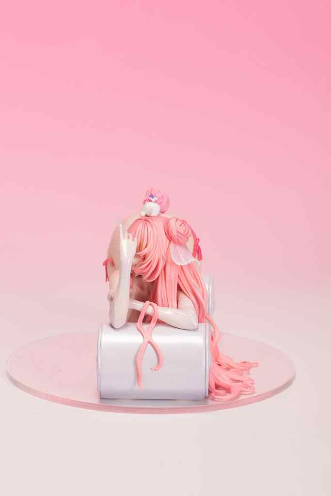A collectible figure of NIKKE ドロシー with long pink hair and detailed features, seated on a silver cylindrical base against a soft pink background.