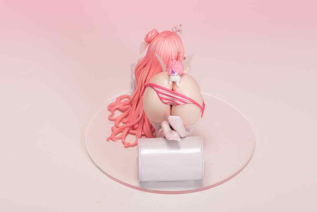 A detailed figurine of a character with pink hair, wearing a revealing outfit, seated on a cylindrical base with a pink background.