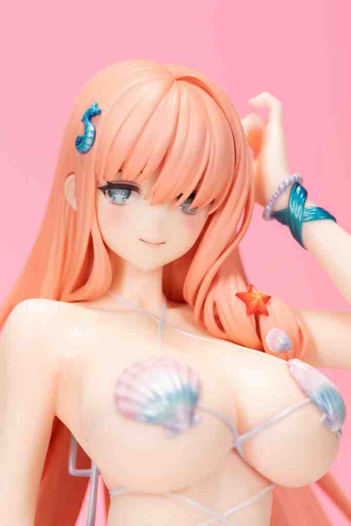 A close-up of a figurine featuring a female character with long, pastel pink hair, decorated with beach-themed accessories, including shells and a starfish, set against a pink background.