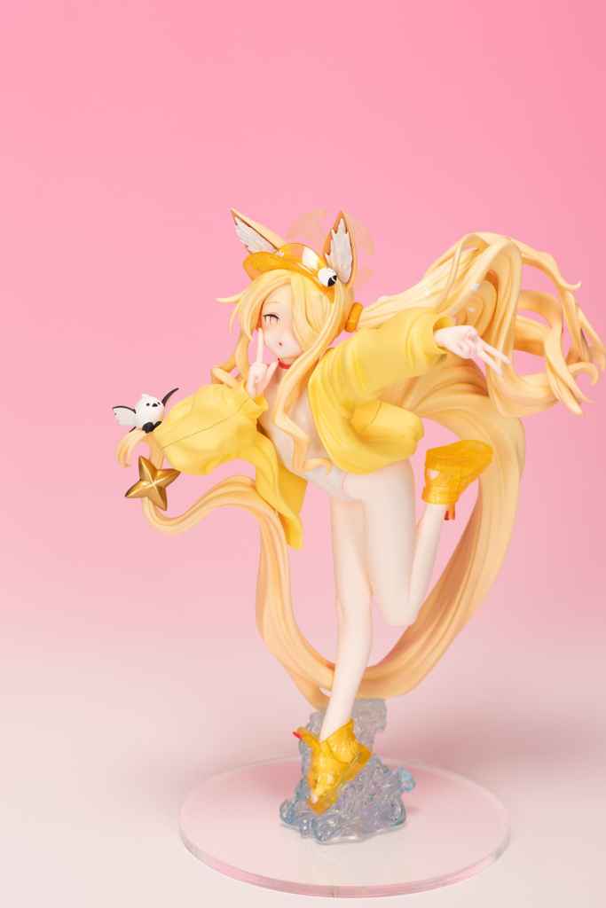 A whimsical figurine of a character with long blonde hair, wearing a bright yellow outfit and cute accessories, posed playfully against a soft pink background.