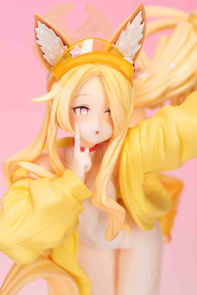 A detailed figure of a character with long blonde hair and cat ears, wearing a yellow oversized jacket, playfully placing a finger to her lips against a pink background.