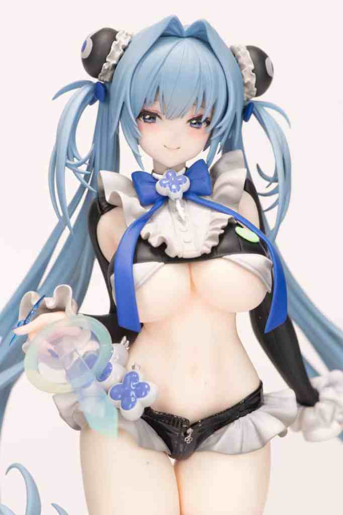 A close-up of a stylized figurine featuring a character with blue hair styled in two pigtails, wearing a black and white outfit with a frilled skirt. The character has a playful expression and is holding a colorful transparent accessory.